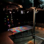 In einem dunklen Raum ist ein Tisch mit einem leuchtenden Bildschirm zu sehen der bunte Lichter und Muster anzeigt. Im Hintergrund sind verschiedene Lichteffekte sichtbar. In einem dunklen Raum ist ein Tisch mit einem leuchtenden Bildschirm zu sehen der bunte Lichter und Muster anzeigt. Im Hintergrund sind verschiedene Lichteffekte sichtbar.