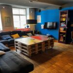 Gemütlicher Wohnbereich mit blauer Wand mit Sofas Holzpaletten-Tisch und einem Fernseher. Helle Fenster und moderner Stil schaffen eine einladende Atmosphäre. Gemütlicher Wohnbereich mit blauer Wand mit Sofas Holzpaletten-Tisch und einem Fernseher. Helle Fenster und moderner Stil schaffen eine einladende Atmosphäre.